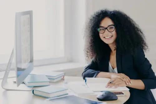 Glad Curly Office Worker Watches Webinar On Business Topic, Writes Down Notes With Pen, Smiles Positively, Wears Spectacles And Black Clothes Prepares Report Wears Transparent Glasses. Coworking Space